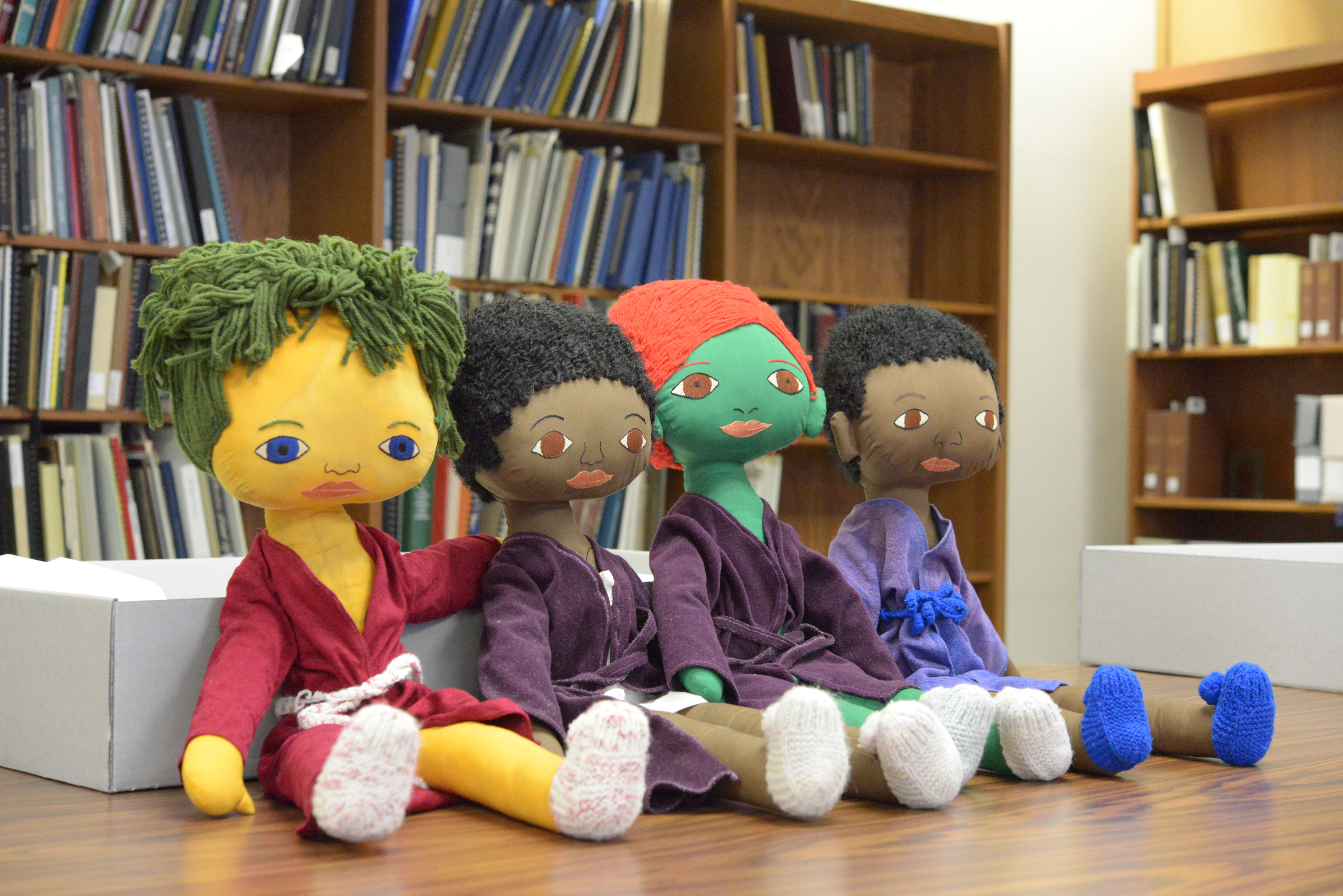 A photo of four cloth dolls of different colors (yellow, brown, green, and brown) wearing robes placed in a row on a table.